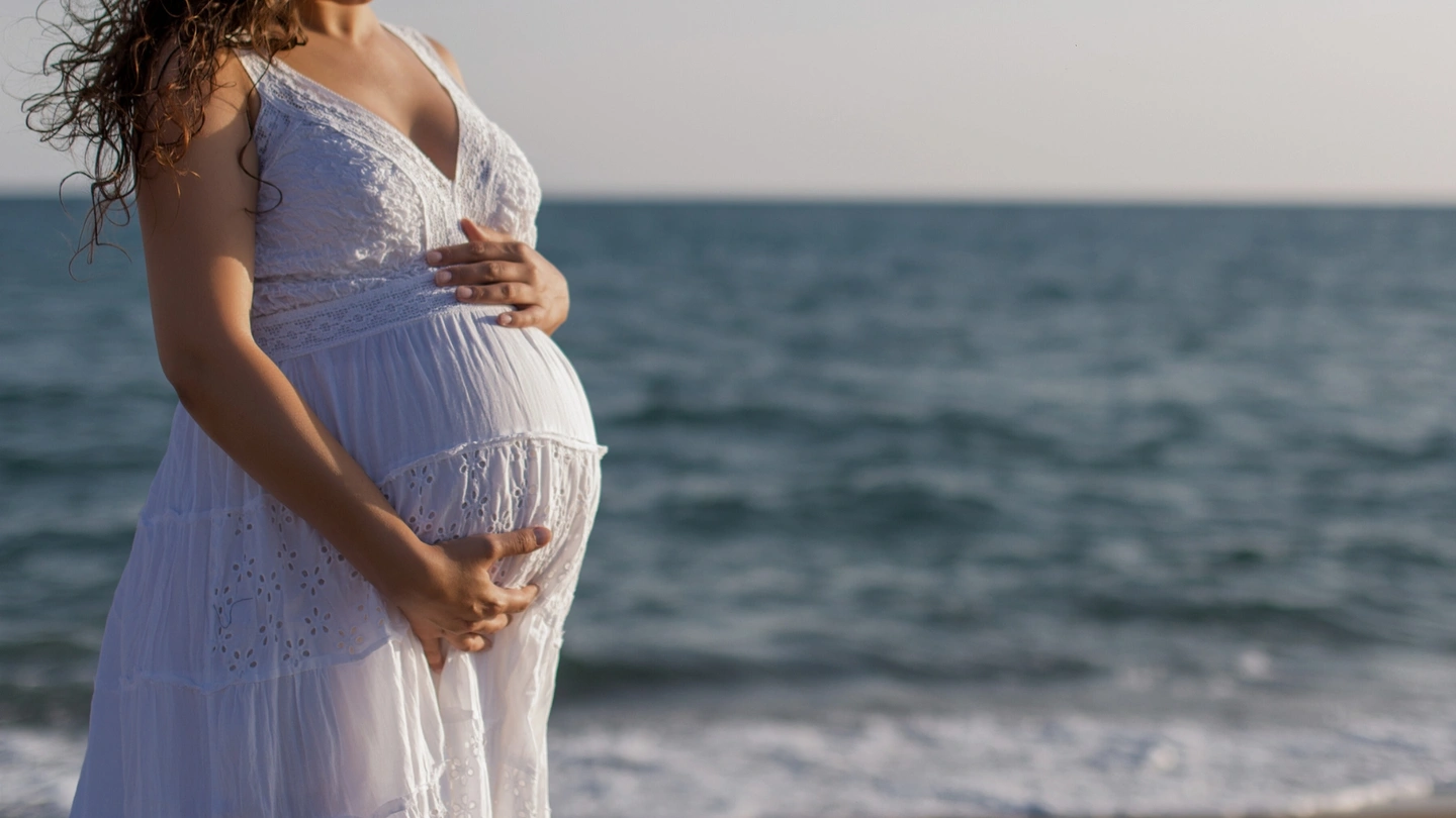 Pregnant woman in a white dress on the beach caressing her belly.